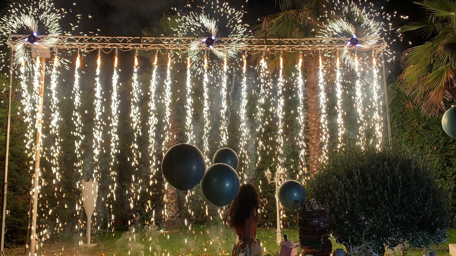 Fuochi d'artificio che cadono a cascata da una struttura metallica dietro palloncini scuri e persone di notte.