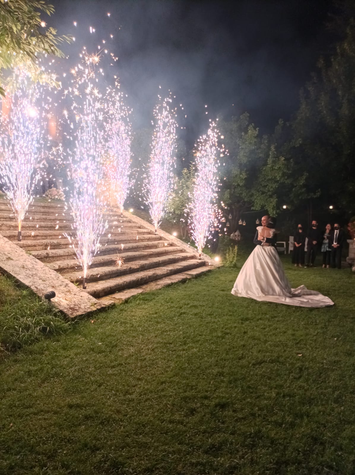 Una sposa in abito bianco è in piedi davanti alle scale, mentre i fuochi d'artificio celebrativi illuminano lo sfondo. Erba verde, alberi e figure scure.