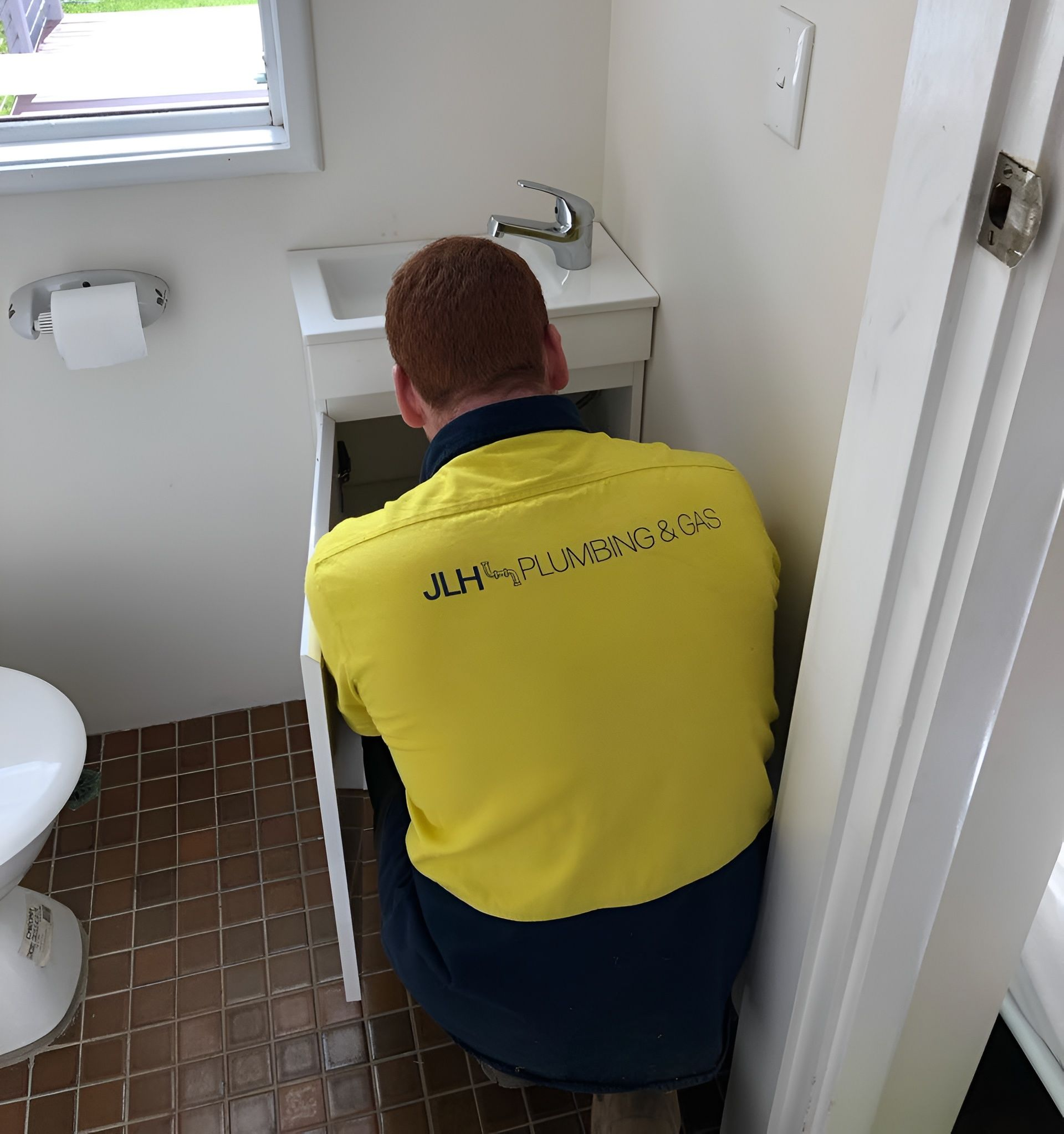 A plumber in a yellow shirt installing a sink in a small bathroom with a toilet — JLH Plumbing, Gas & Roofing in South Nowra, NSW
