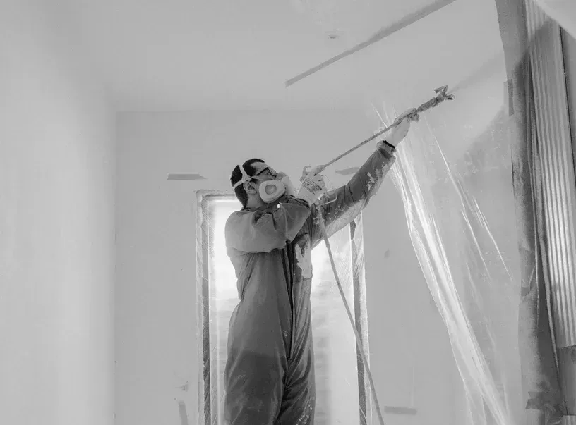 Un operaio con indumenti protettivi vernicia a spruzzo una stanza con finestre e pareti coperte da teli di plastica.