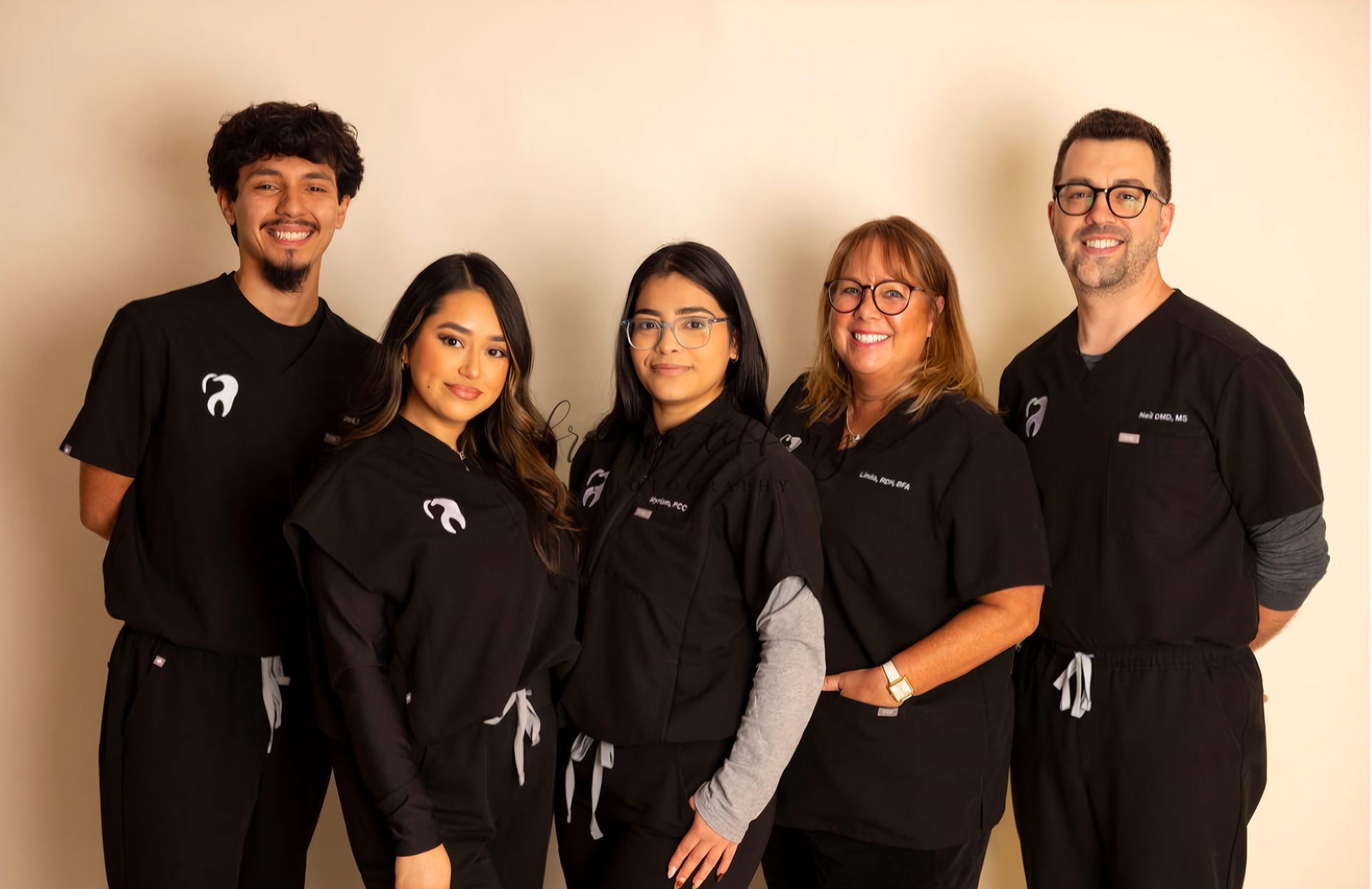 Dental team in black scrubs, smiling, posing for a group photo. Light background.