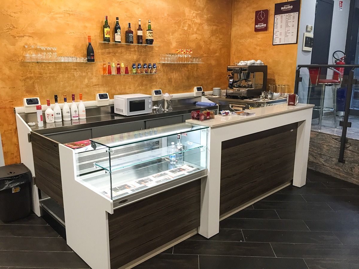 Small café counter with pastry display, coffee machine, and bottles on a warm orange wall