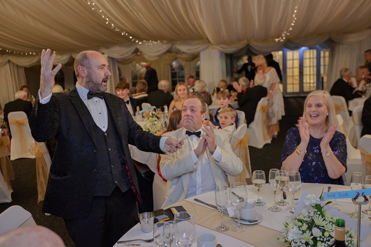 A man in a tuxedo is standing in front of a crowd of people at a wedding reception.