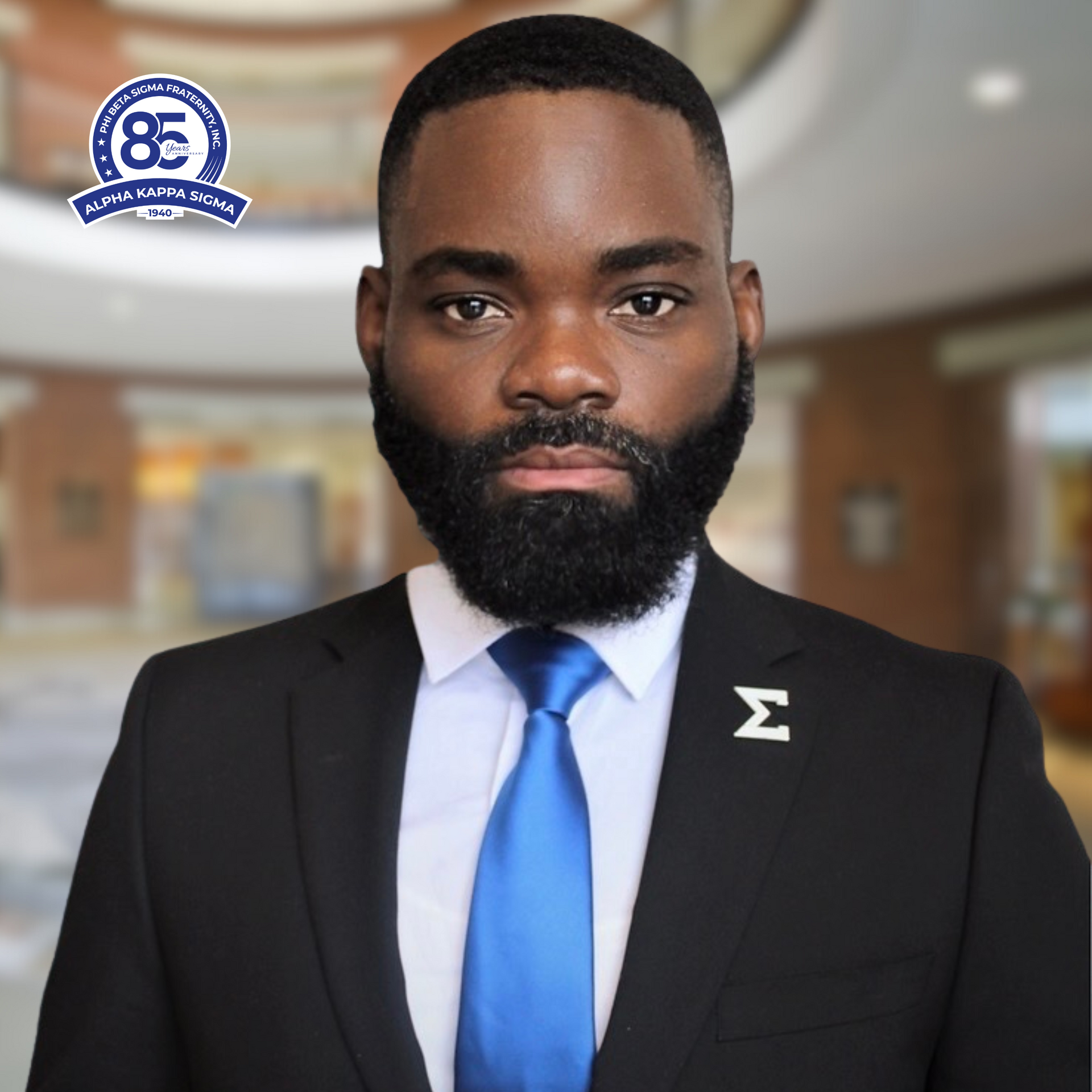Man in suit, blue tie, black beard, Sigma emblem, and a blurred background.