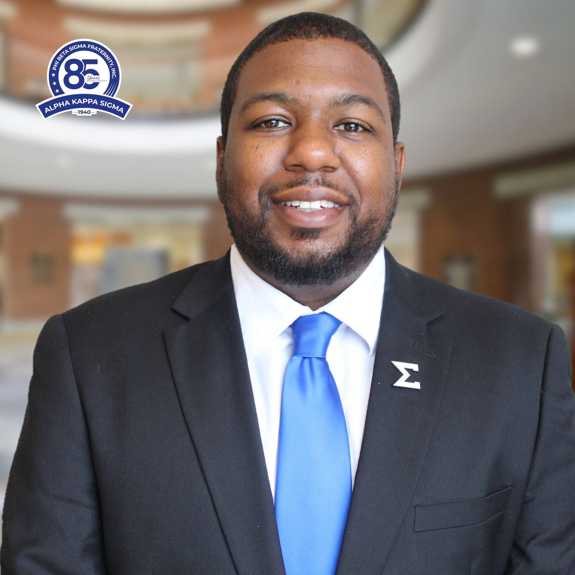 Man in black suit and blue tie, smiling; Sigma logo pin.