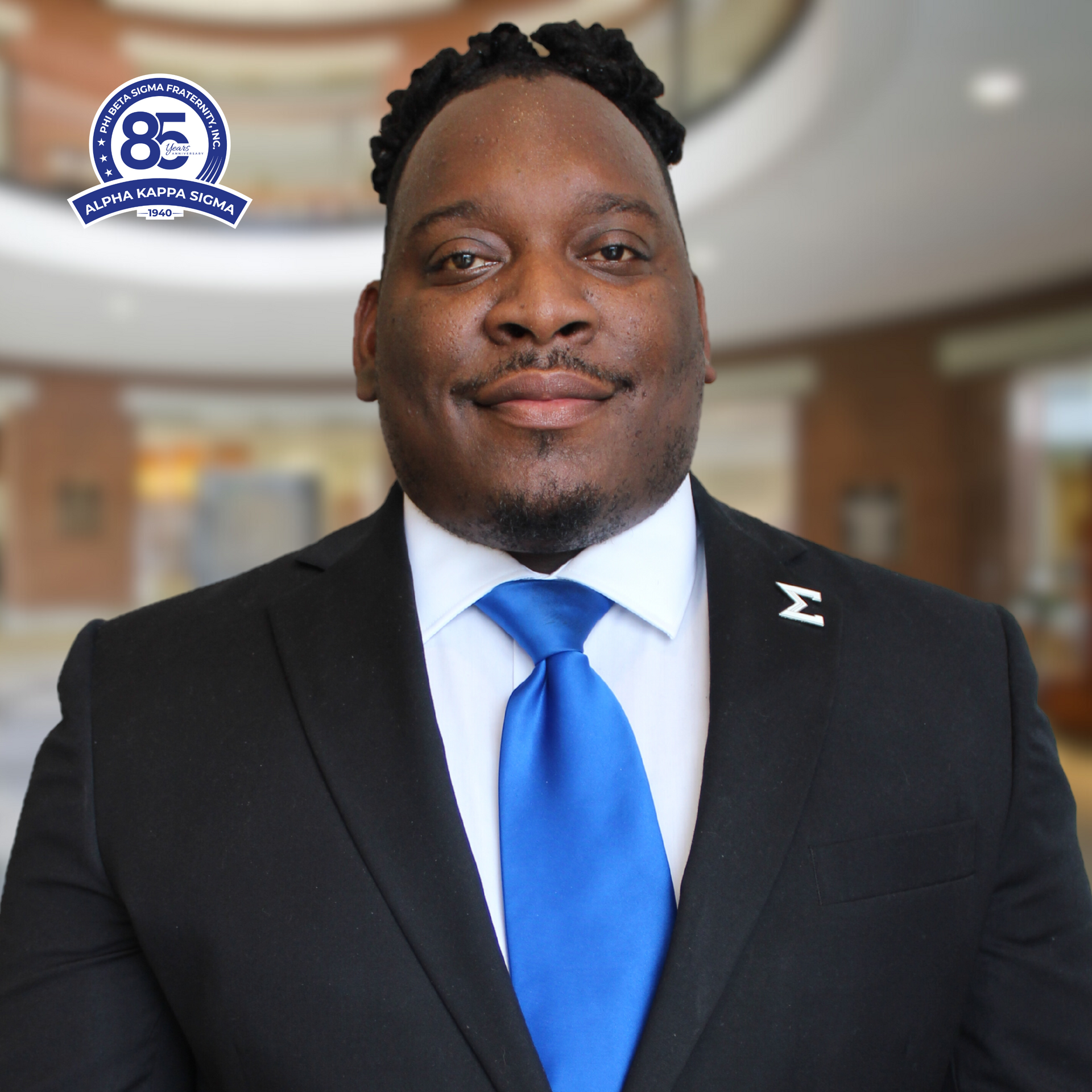 Man in a suit with a blue tie, standing in a building. He wears a fraternity pin.