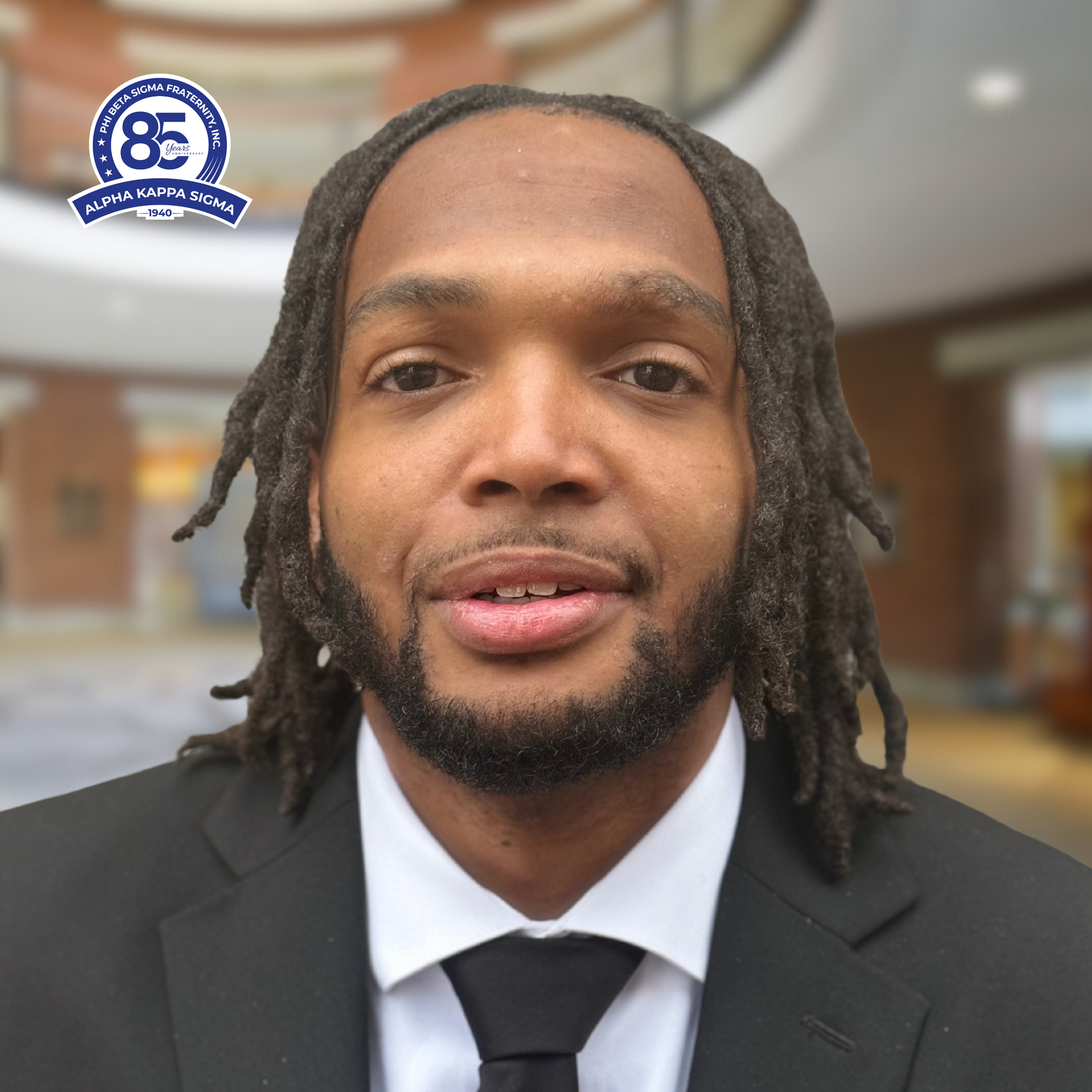 Man with dreadlocks in black suit and tie, smiling, indoors.