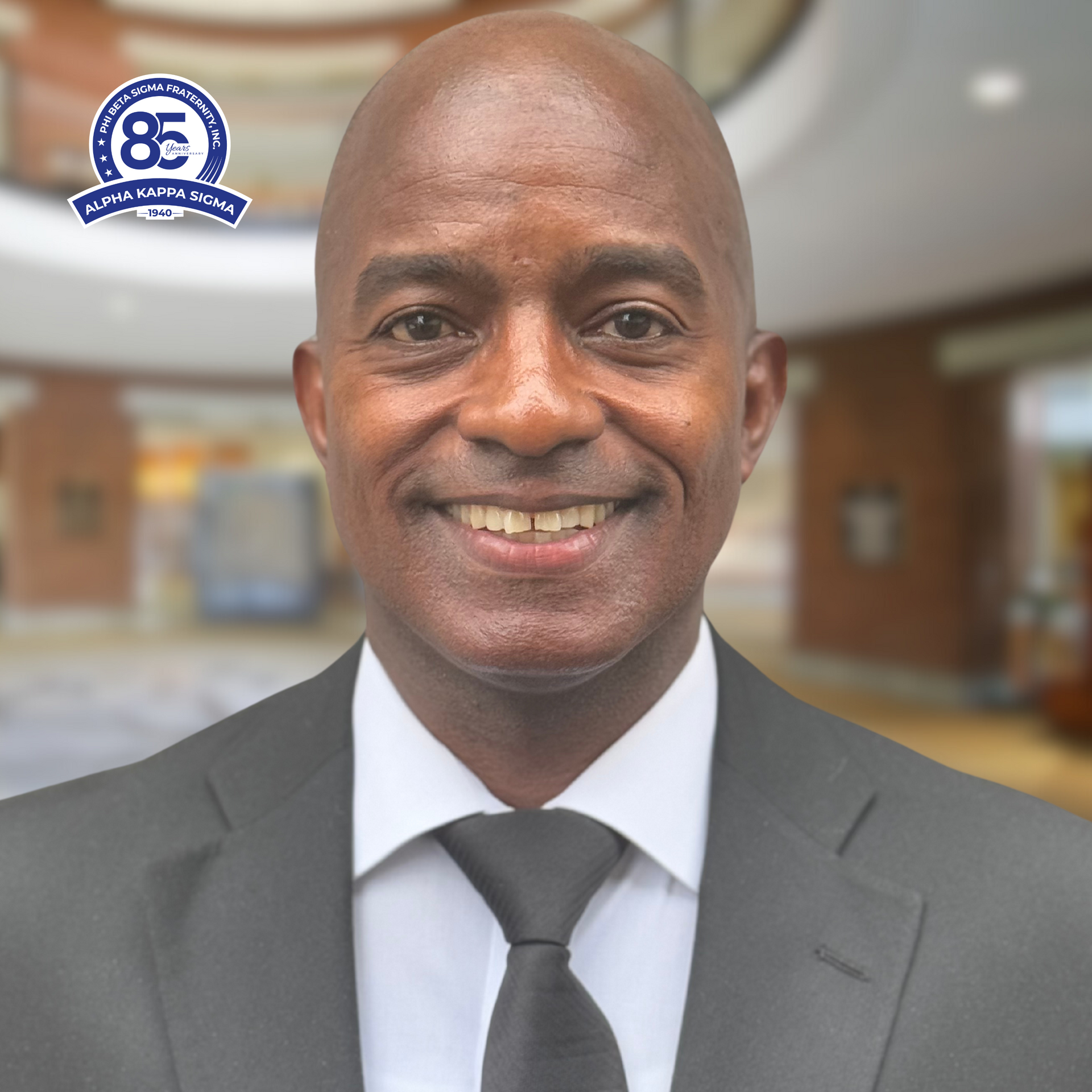 Man in a suit smiles, with a background of a building lobby, a badge on the top left.