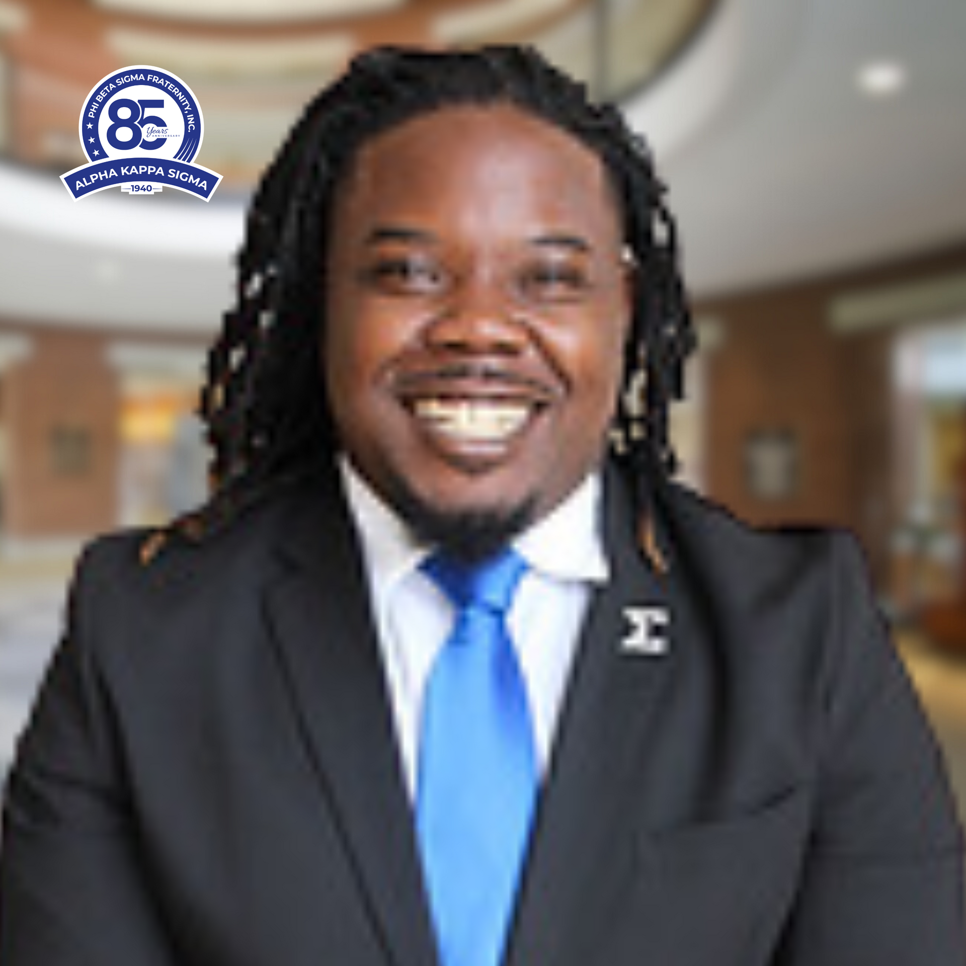 Man in suit with a blue tie, smiling, indoors with a logo in the background.