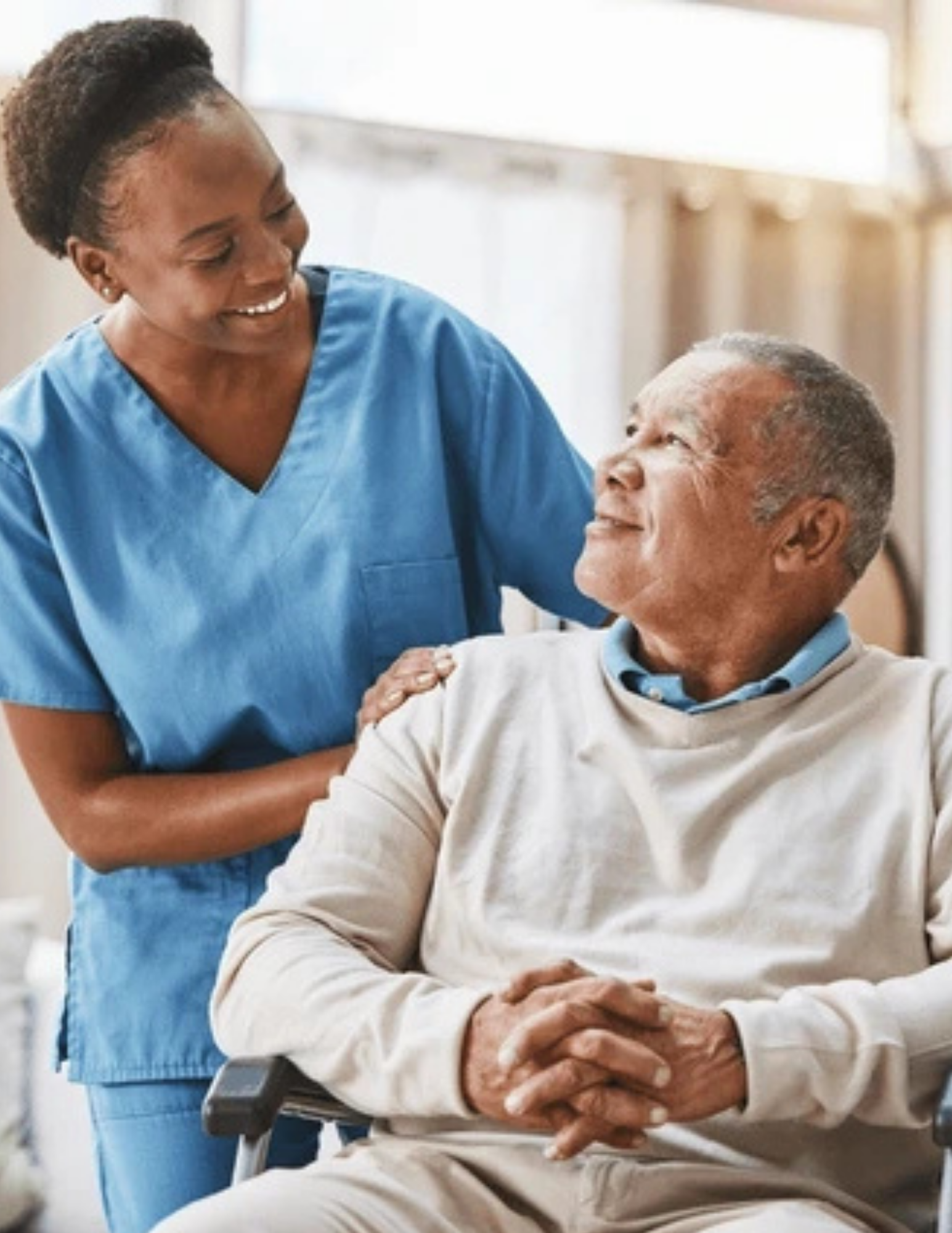 Caregiver in blue scrubs smiles at a man in a wheelchair; they look at each other with affection in a bright room.