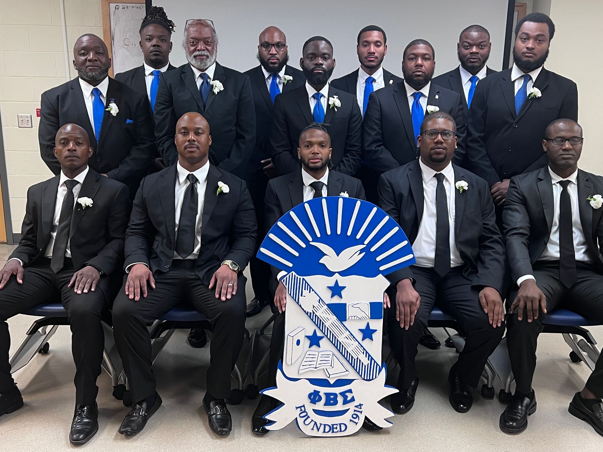 Alpha Kappa Sigma Chapter. Group of men in black suits with blue ties, posing with a fraternity crest.