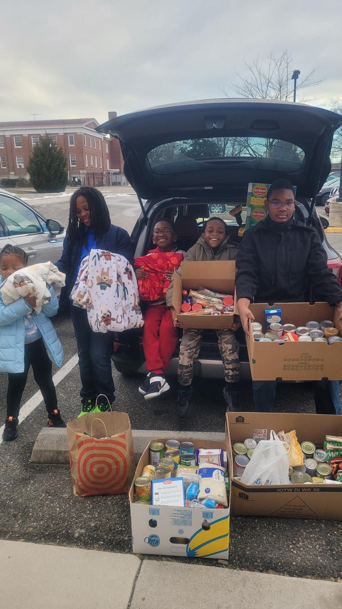 People unloading canned goods and clothing from a car trunk in a parking lot.