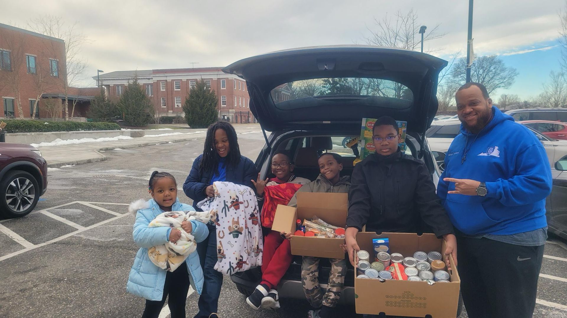People unloading donations from a car in a parking lot. Two adults and three children hold boxes and clothes.