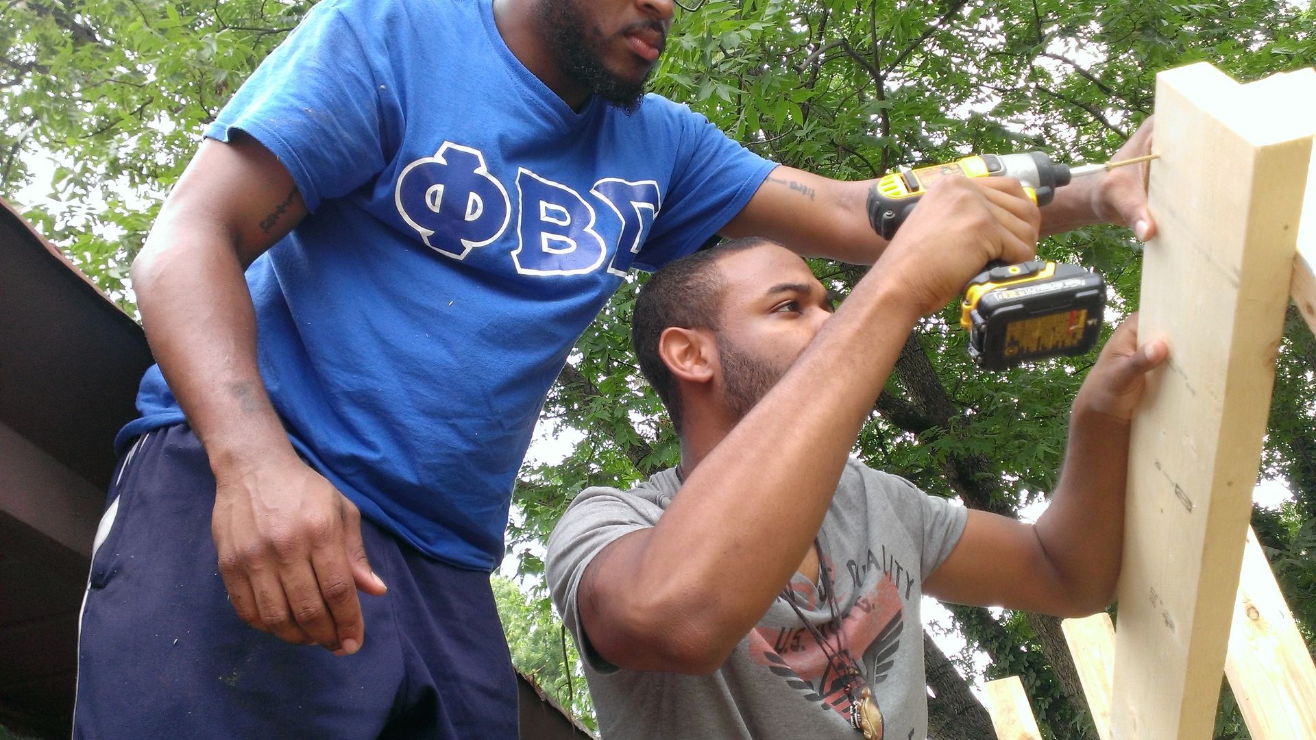 Two men using a power drill to assemble wooden planks outdoors. One wears a blue shirt, the other a gray shirt.