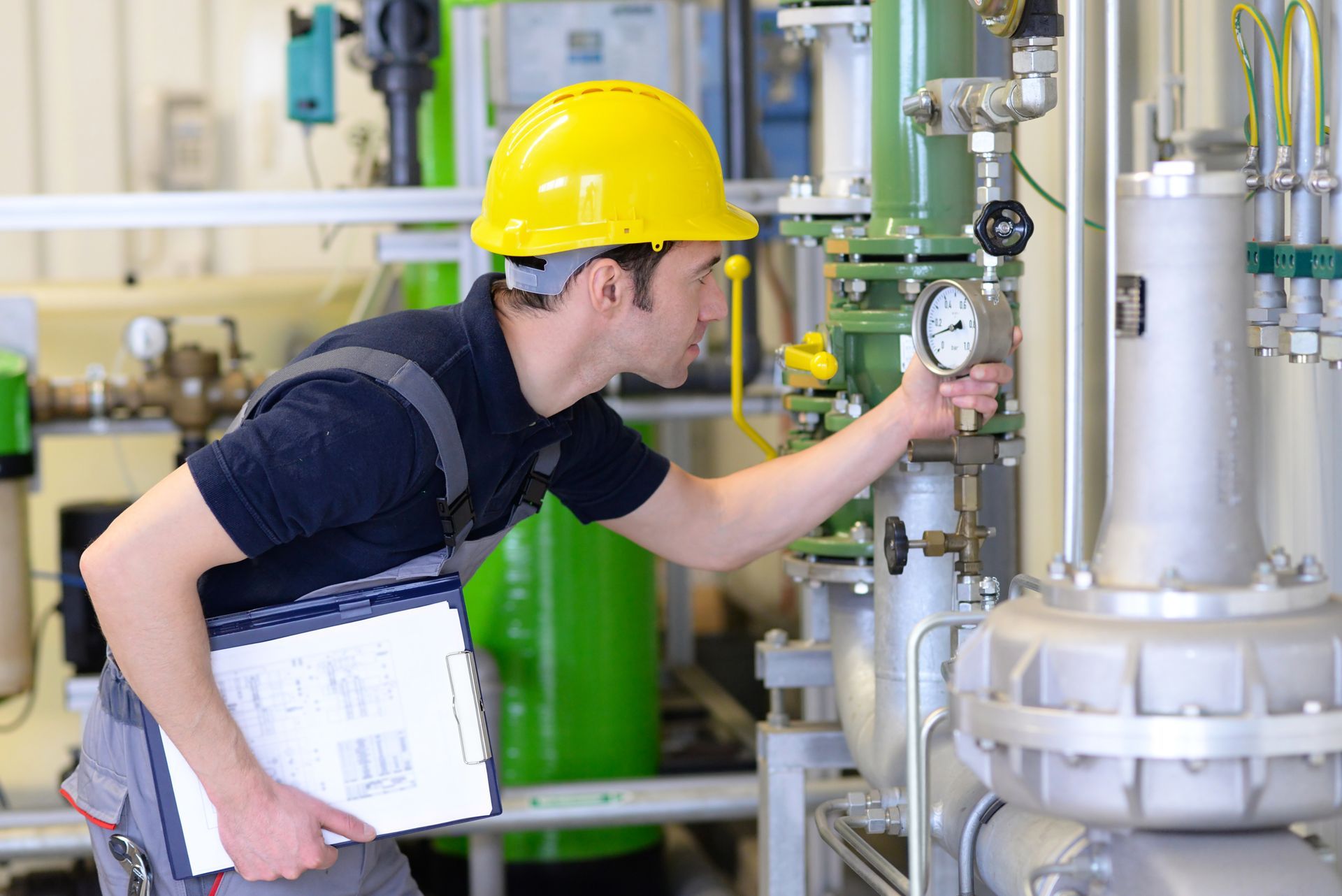 Eine Person mit gelbem Schutzhelm überprüft die Messgeräte an Industriemaschinen. Sie hält ein Klemmbrett.