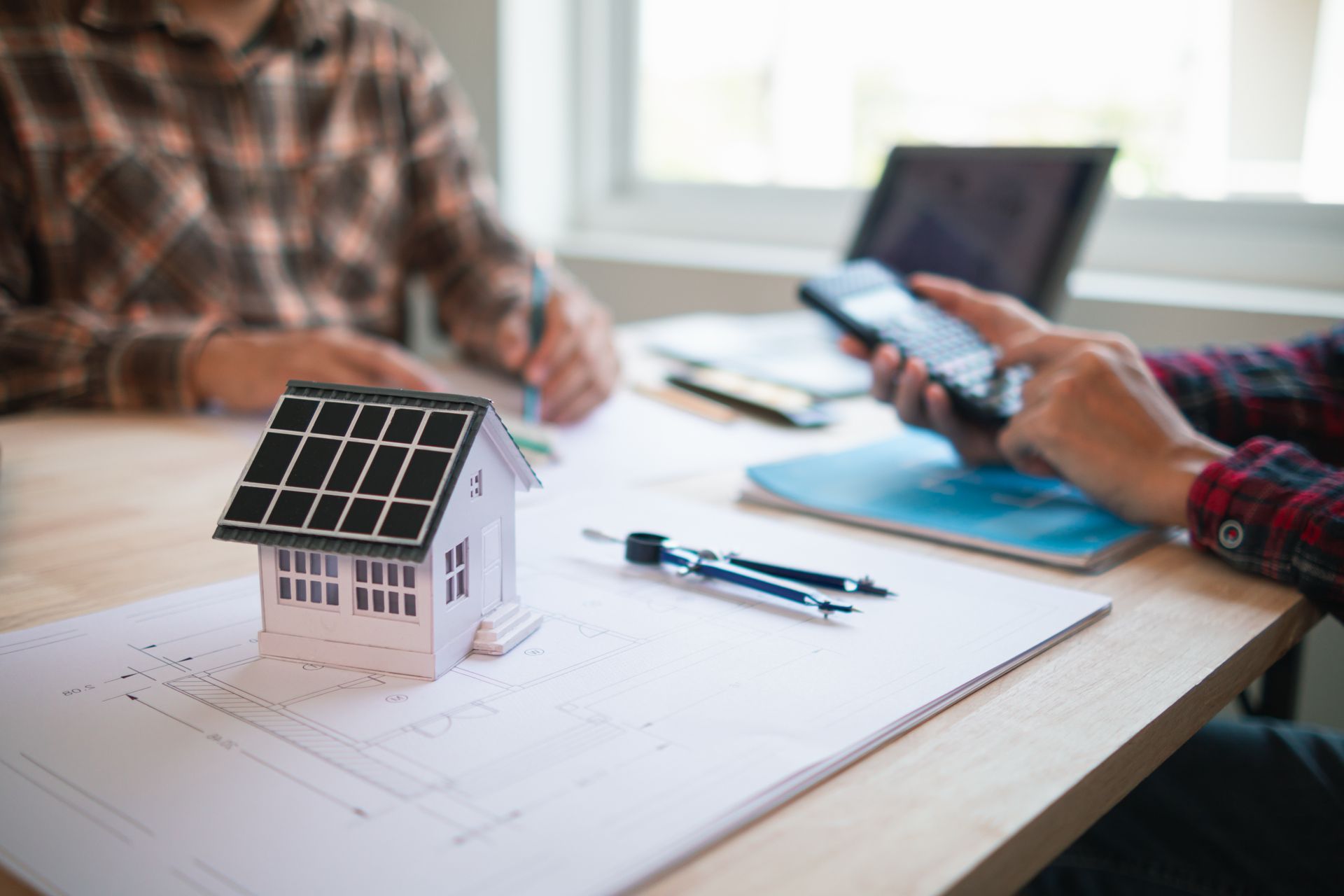 Miniaturhaus mit Solarpaneelen auf einem Bauplan, zwei Personen an einem Tisch, die Pläne durchgehen