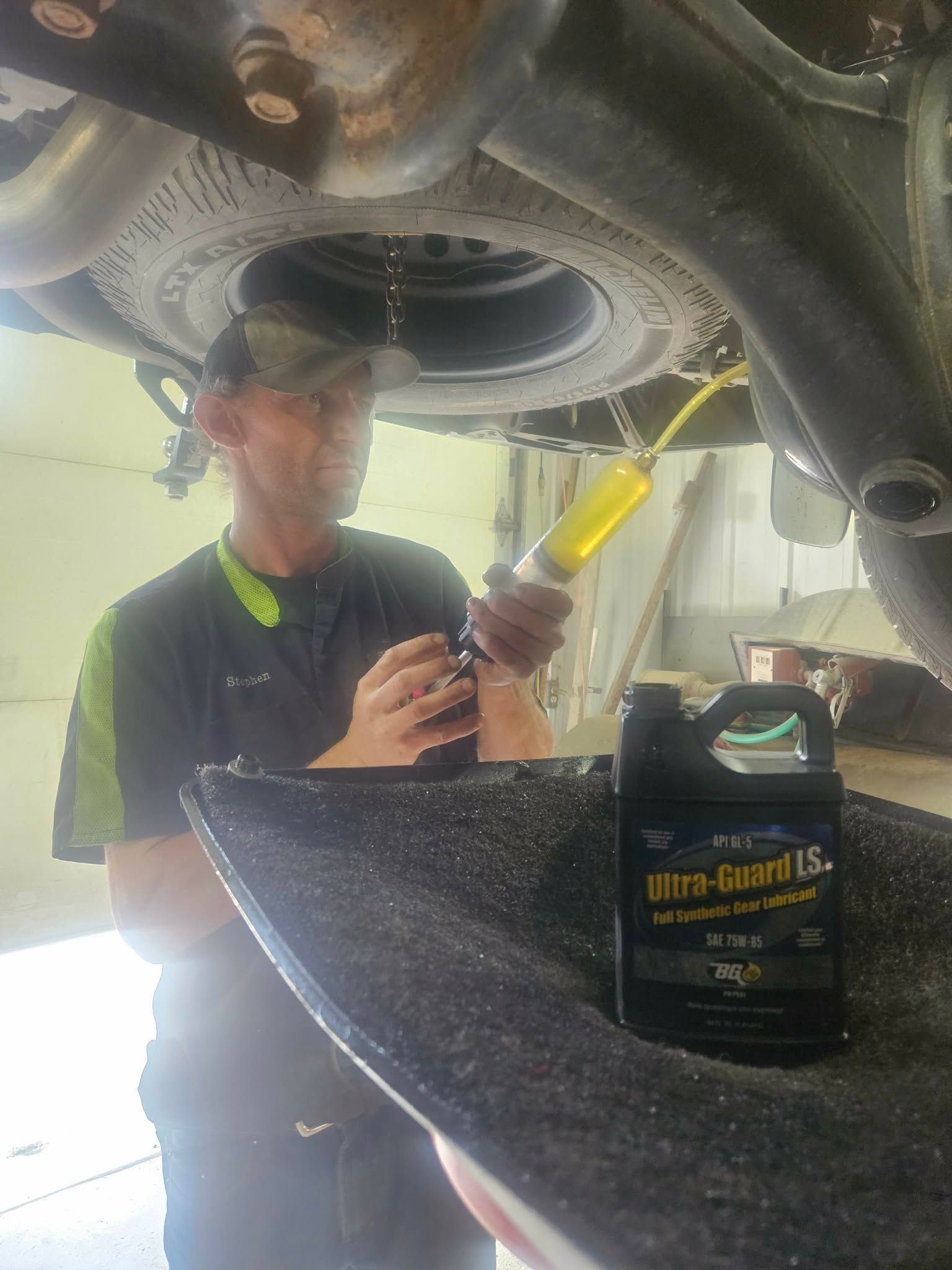 Mechanic under a car filling a fluid reservoir with a yellow funnel; oil container in foreground.