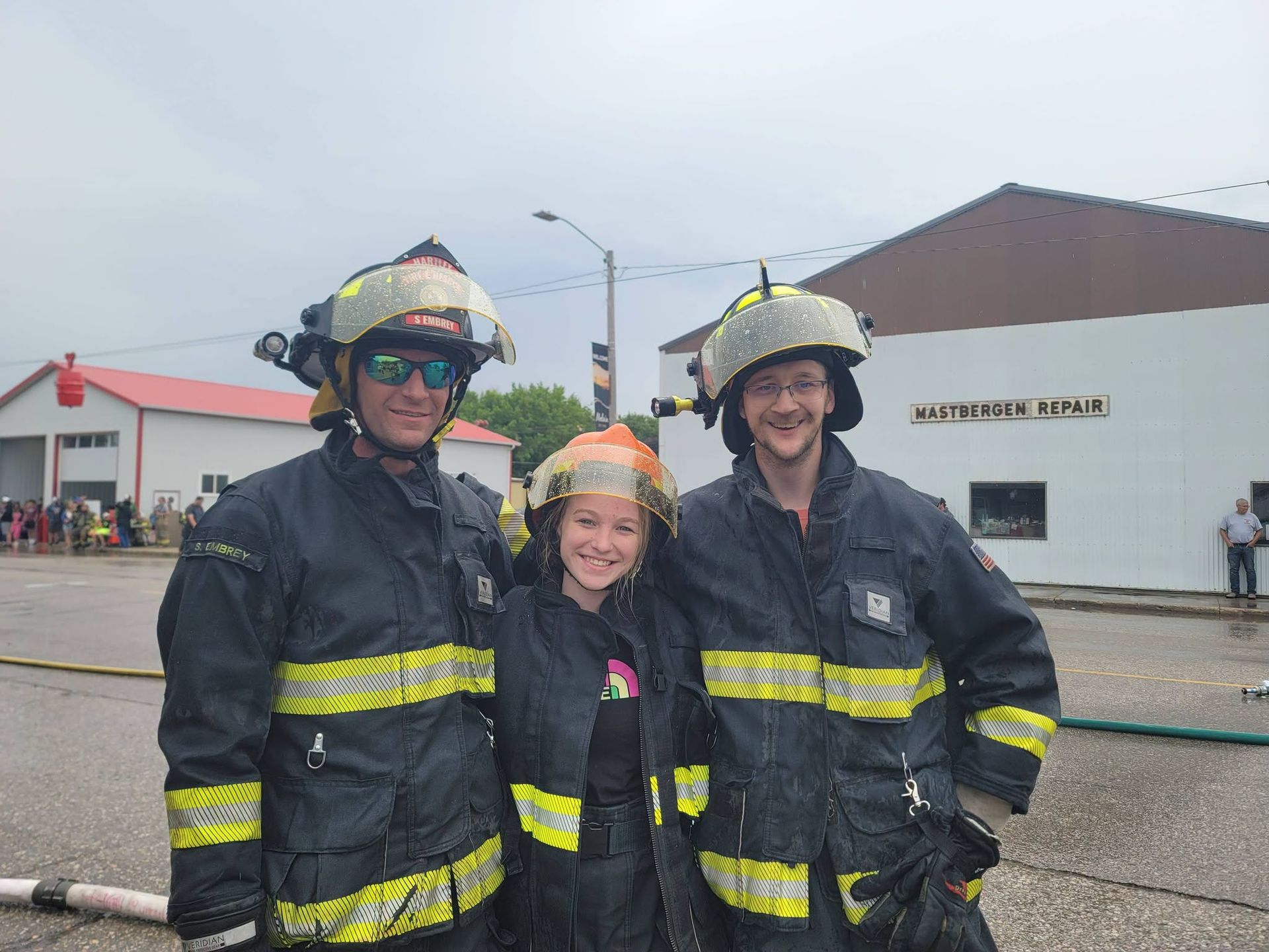 Three firefighters in black turnout gear and helmets stand smiling in front of a fire station.