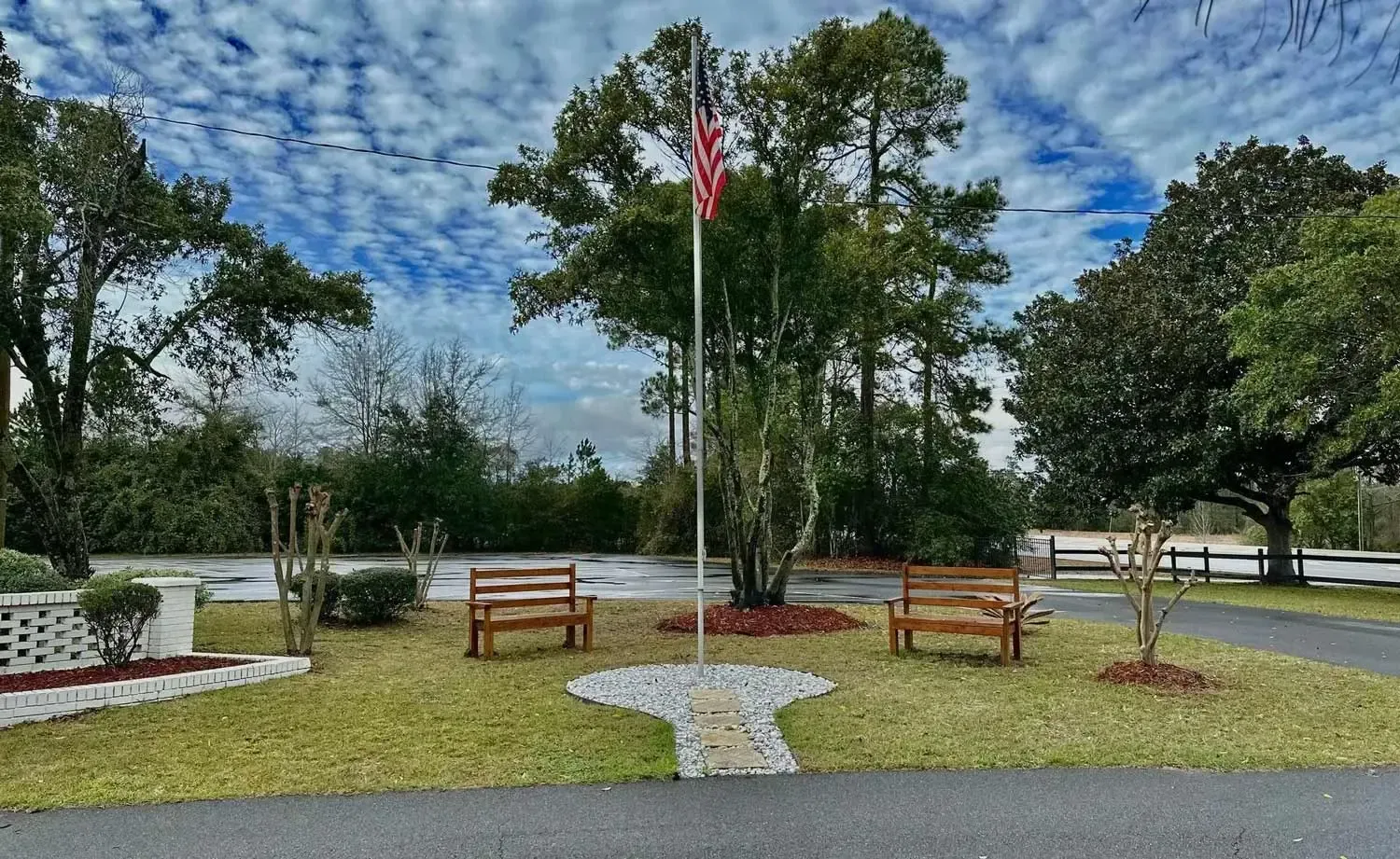 Park memorial with two wooden benches, flagpole, and stone walkway under a cloudy blue sky.