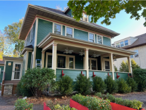 Two-story green house with a porch and red accents, set against a blue sky.