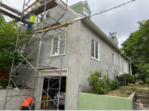 House exterior with scaffolding; workers plastering. Garage open below. Gray, green, and white. Overcast sky.