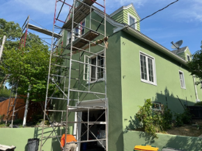 Green house with scaffolding, worker, and tools outside, in daylight.