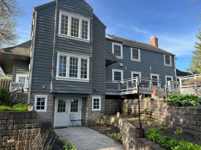 Gray wooden house with white-framed windows, walkway, and stone retaining walls under a blue sky.