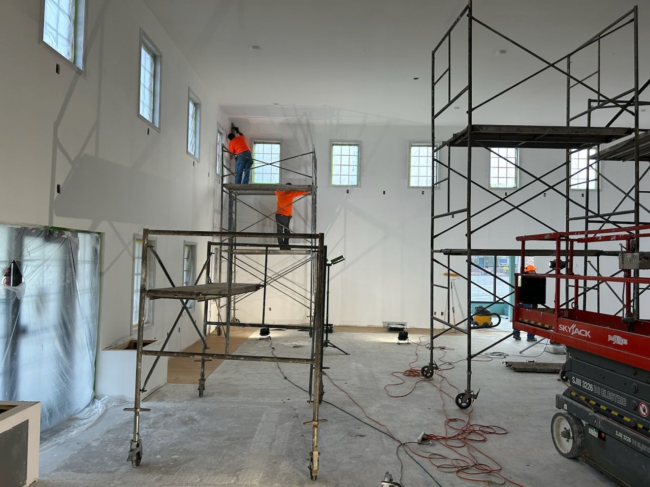 Construction workers on scaffolding inside a building; unfinished walls, windows, and tools present.