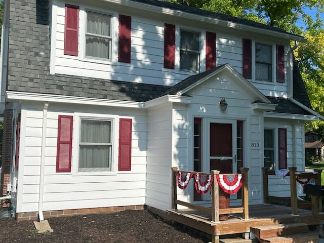 White two-story house with red shutters and door, small porch with patriotic bunting, sunny day.