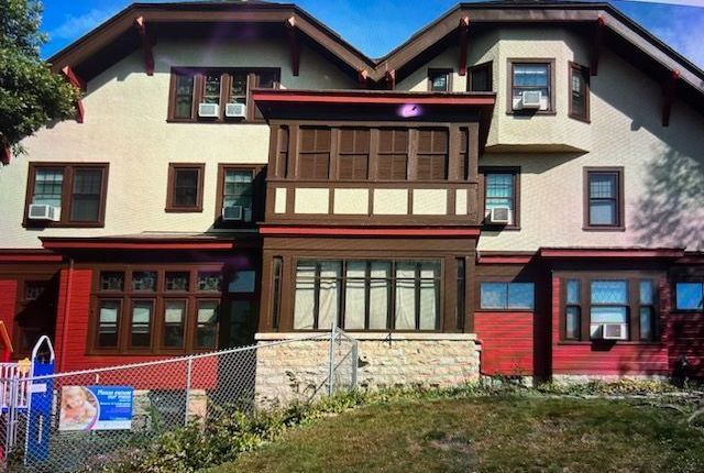 Two-story building with red and tan exterior. Front porch has brown trim and a stone base. A chain-link fence is in the foreground.