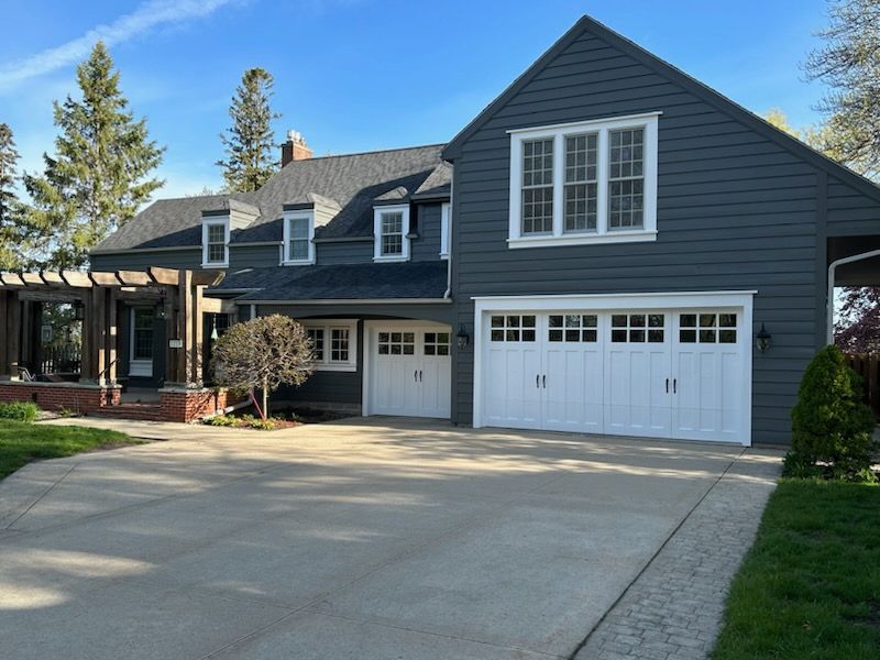 Two-story house with dark gray siding, white garage doors, and a concrete driveway on a sunny day.