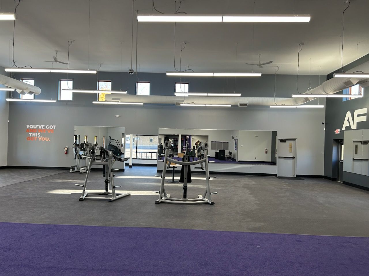 Gym interior with weight machines, mirrors, and purple and gray flooring. Fluorescent lights and windows are present.