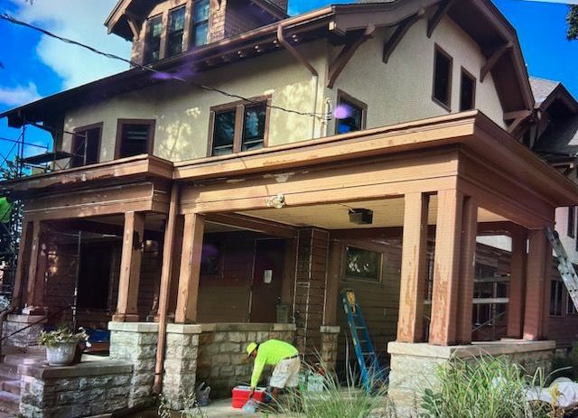 Two-story Craftsman home with brown trim and tan siding. A person in green works on the porch.