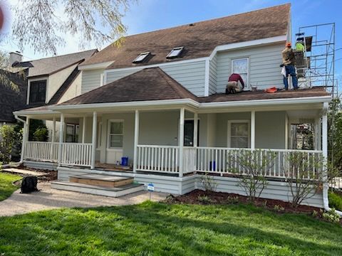 House exterior with workers on roof and porch. Light blue siding, white trim, brown roof.
