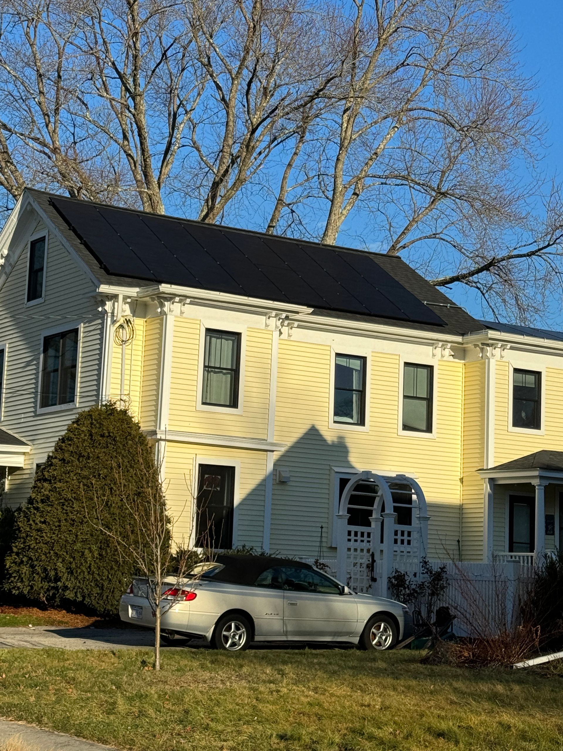 Yellow two-story house with black roof and parked silver car in front under bare trees