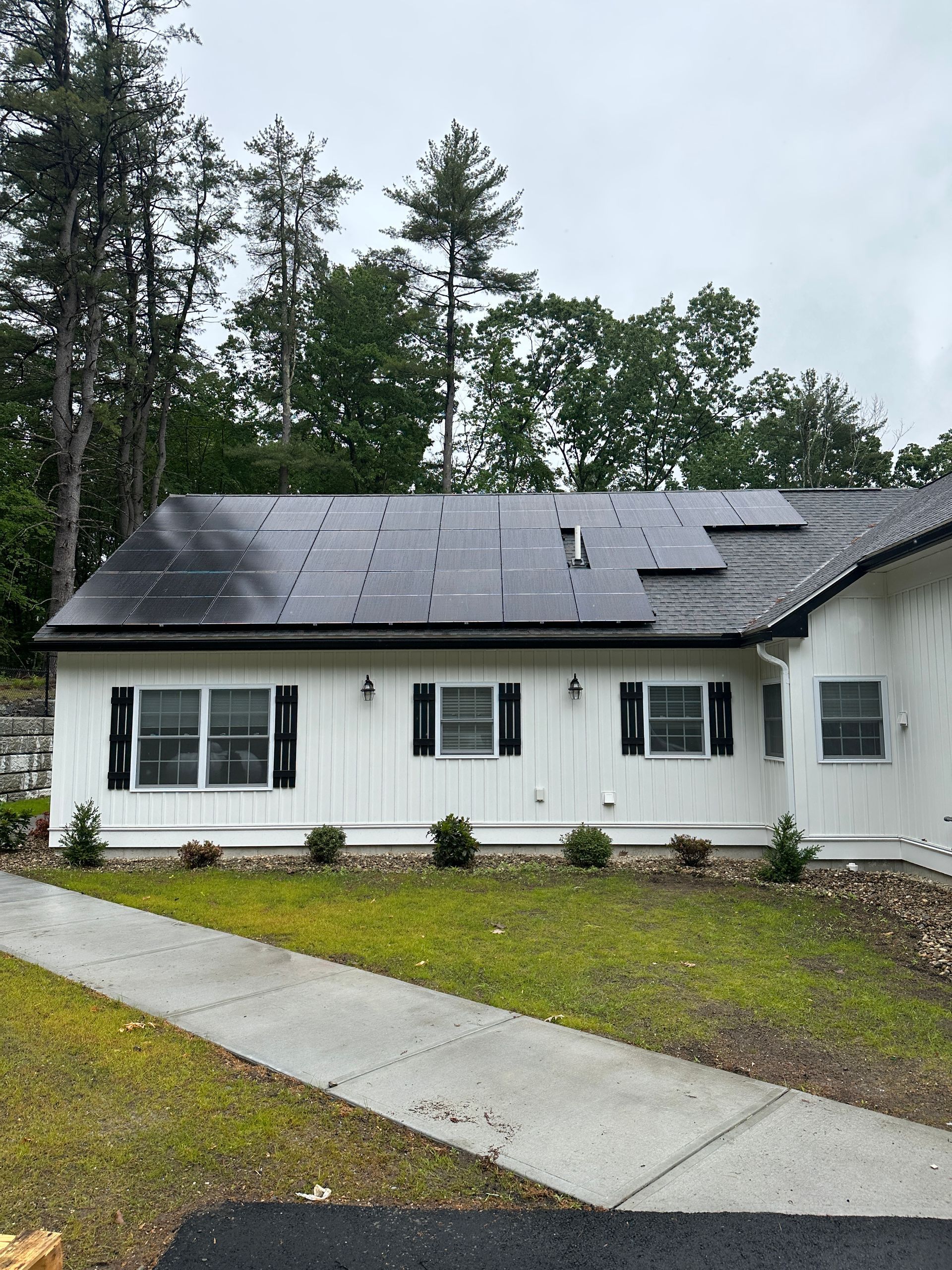 White single-story house with black roof, front windows, and a wet concrete driveway on a rainy day