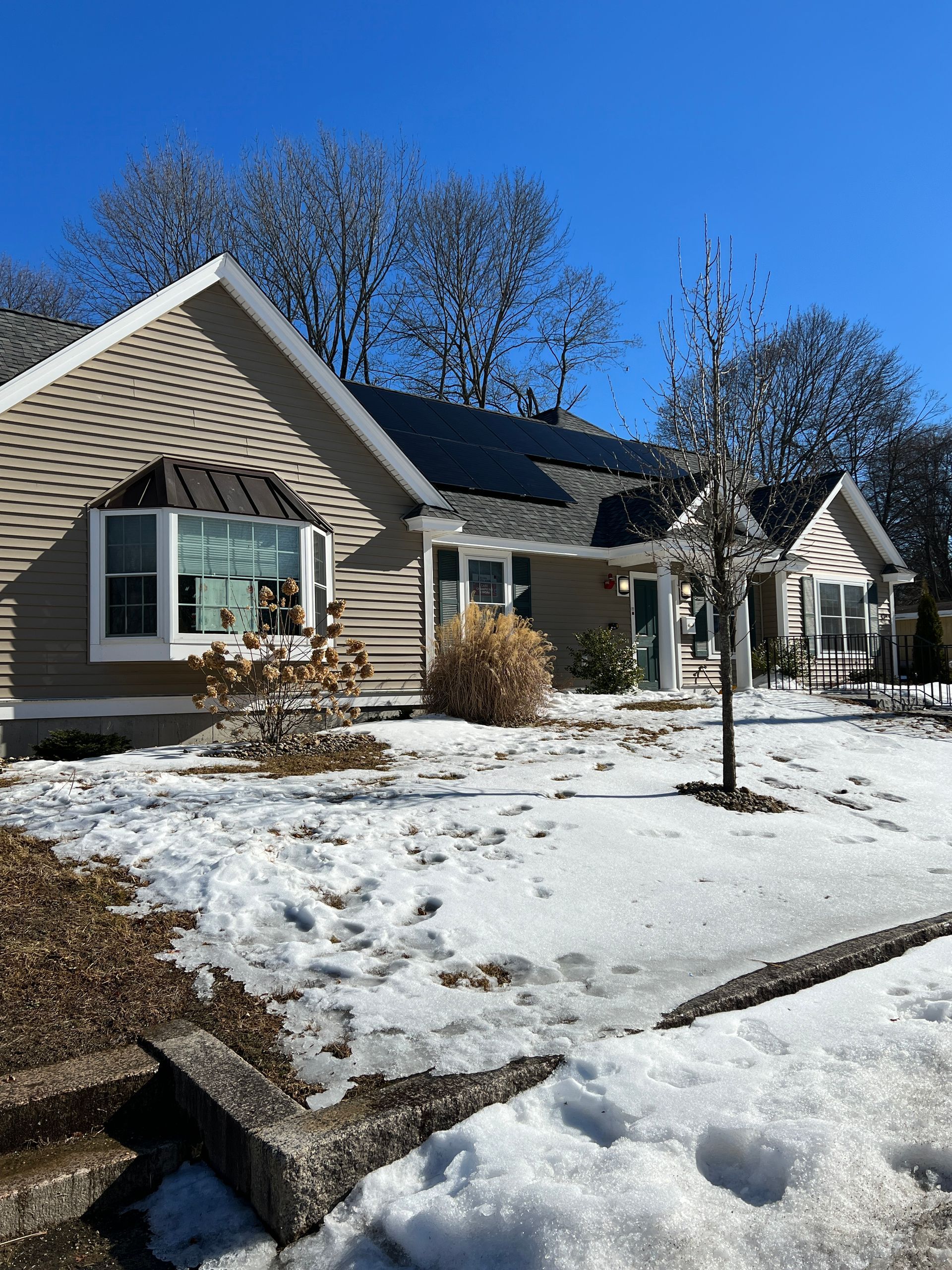 Snow-covered suburban house with beige siding, black roof, and a clear blue sky.