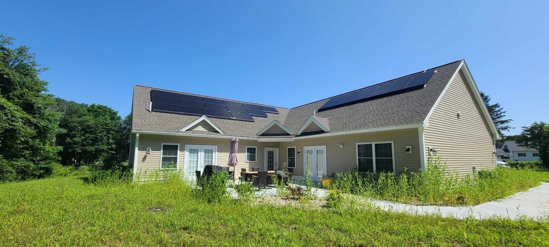 Single-story beige house with dark roof, white trim, and overgrown green lawn under a bright blue sky