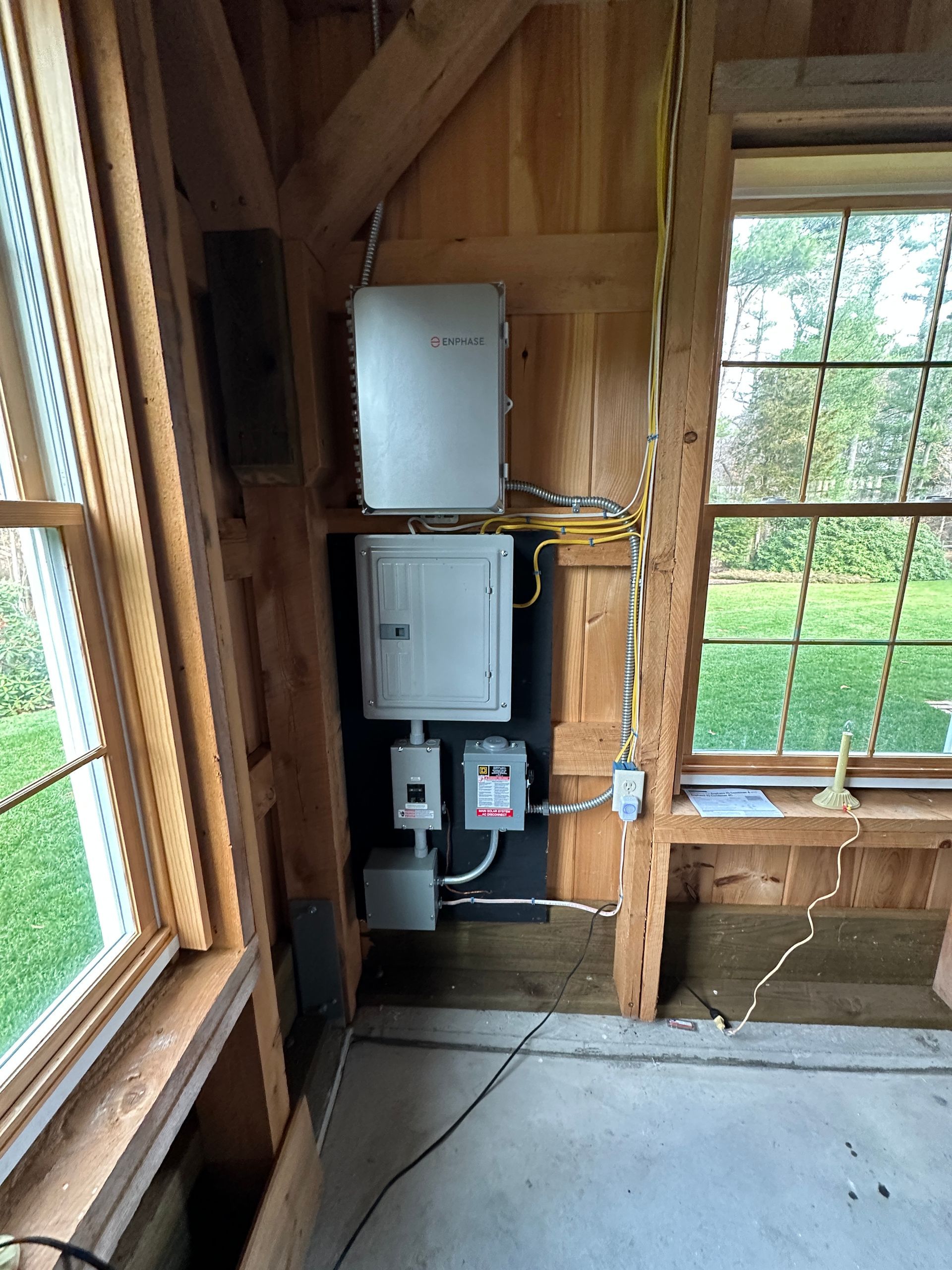 Electrical boxes and wiring mounted on a wood wall inside an unfinished room with windows.