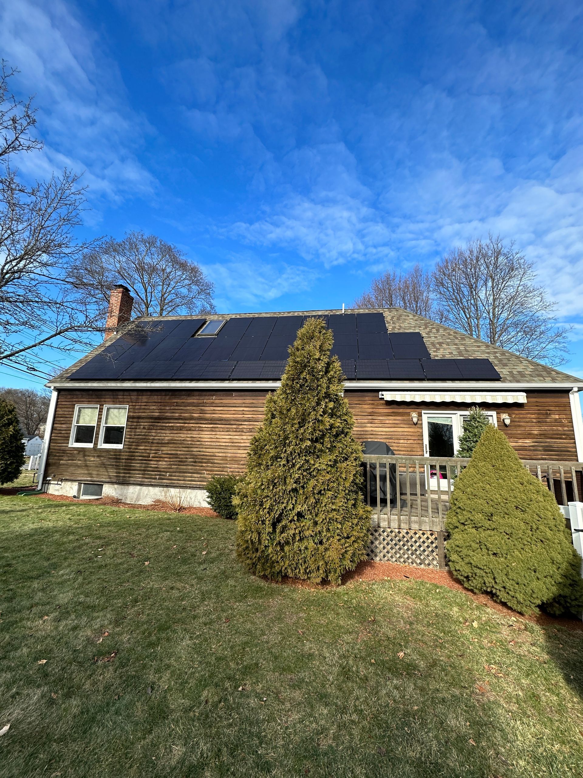 Wooden house with solar panels, a chimney, and two evergreen shrubs under a bright blue sky
