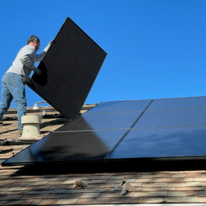 Worker installing black solar panels on a shingled roof under a clear blue sky
