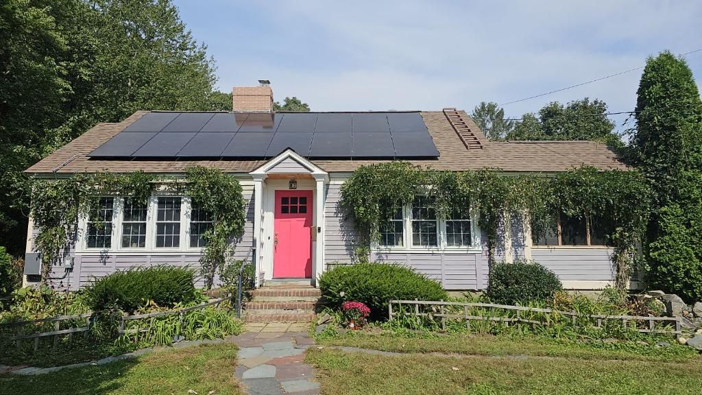 Small white house with a pink front door, solar panels on the roof, and a garden in front