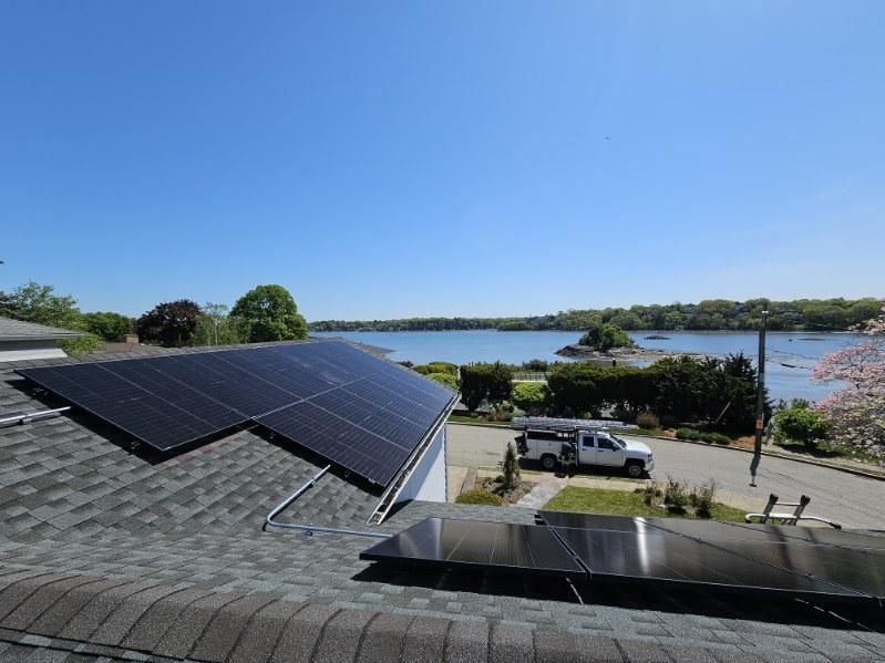 Rooftop solar panels overlooking a sunny waterfront with parked cars and trees.