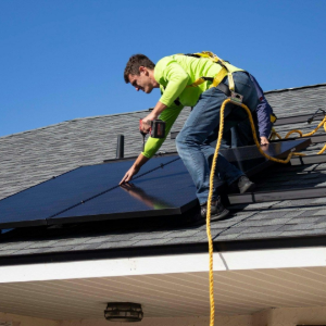 Worker installing solar panels on a roof, with tools and safety rope visible.