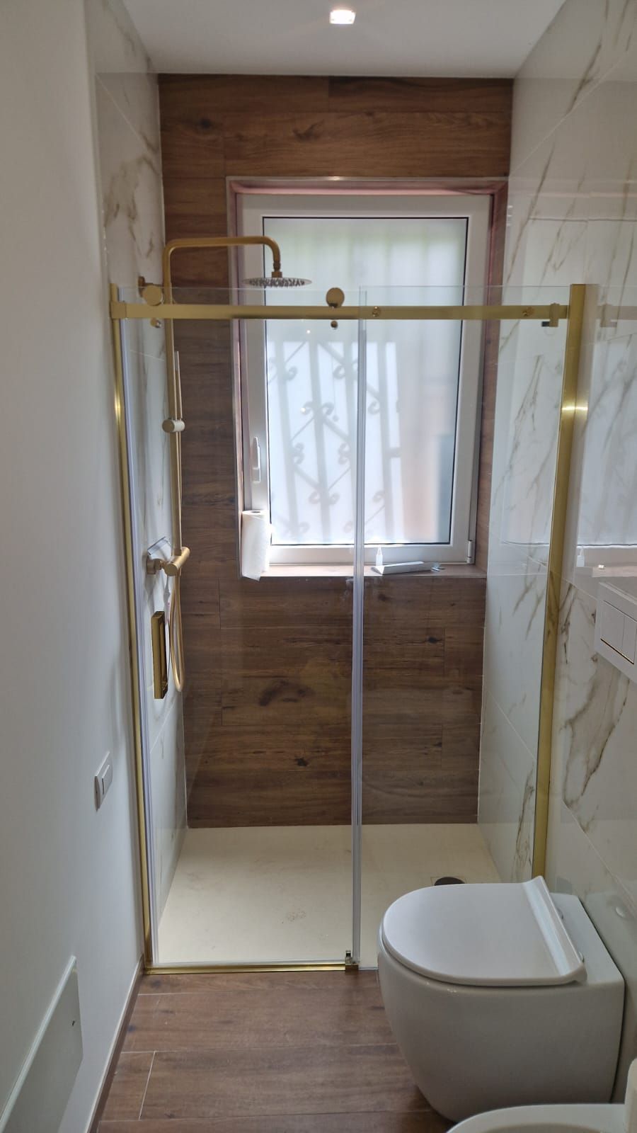 Bagno stretto e moderno con cabina doccia, finestra in vetro, rubinetteria dorata e WC sospeso.