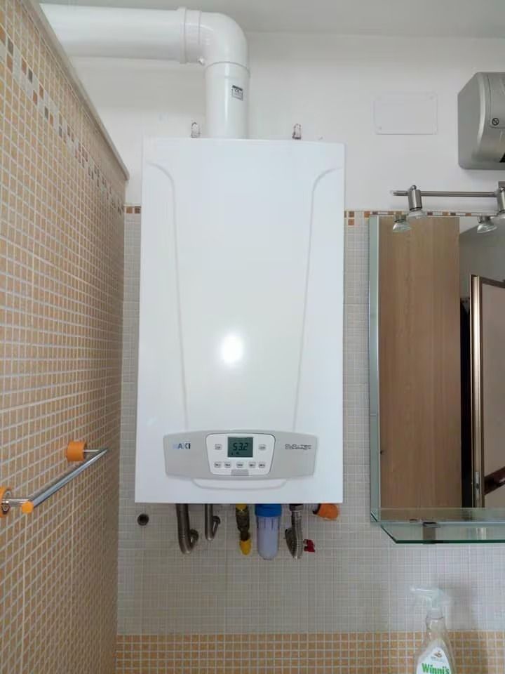 Caldaia bianca a parete in un bagno piastrellato, con tubature e un piccolo pannello di controllo.