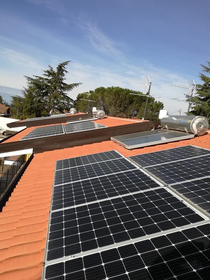 Pannelli solari su un tetto di tegole rosse sotto un cielo azzurro, con alberi e case vicine in vista.