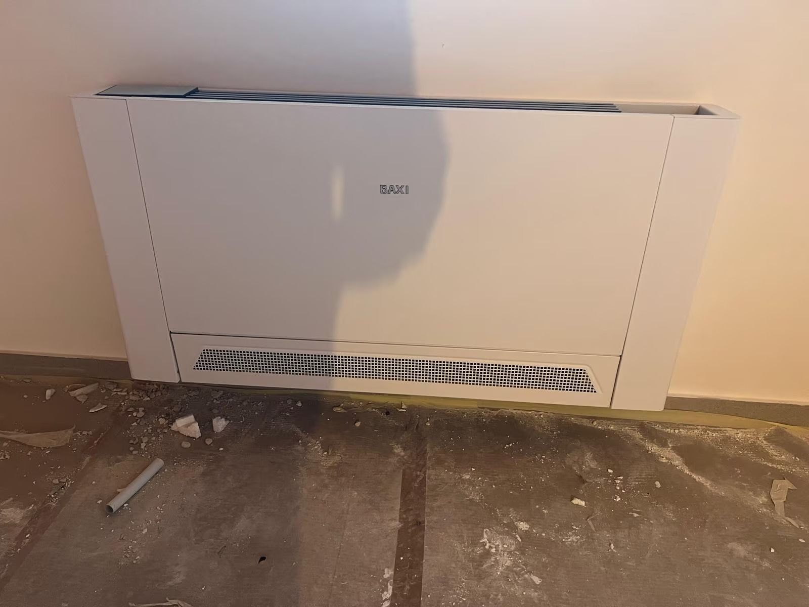 Copertura bianca per termosifone a parete in una stanza polverosa con pannello di ventilazione inferiore danneggiato.