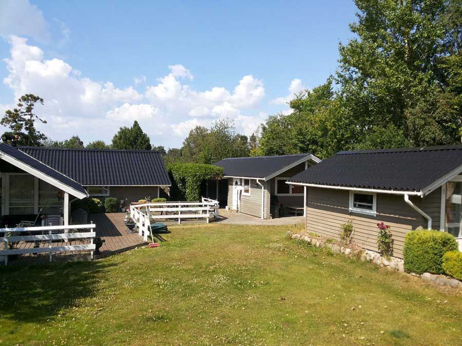 Flere små hytter med mørke tage på en græsklædt plæne under en blå himmel med træer.