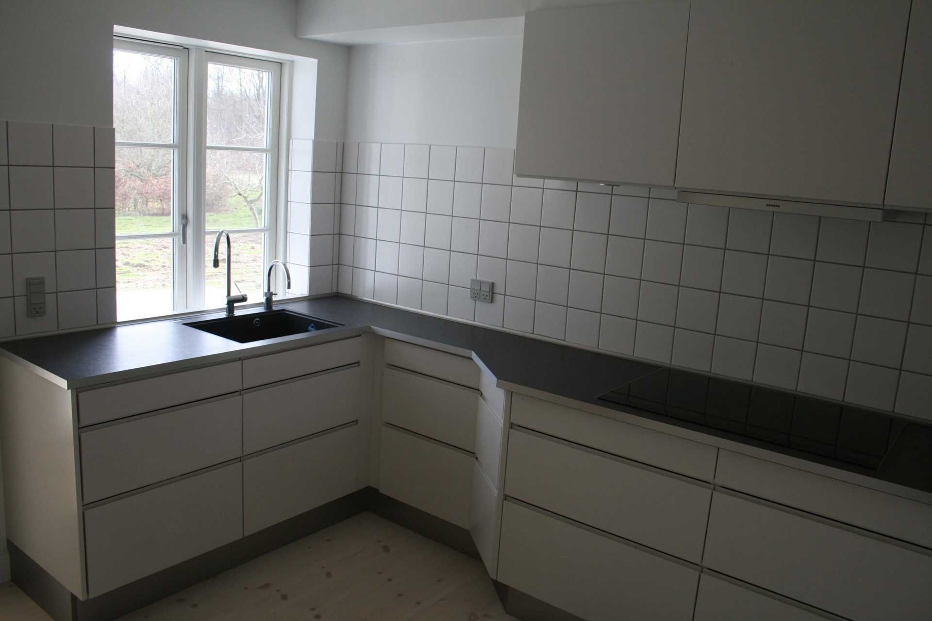 Hvidt køkken med mørke bordplader, et vindue og firkantet hvid flisebagplade.